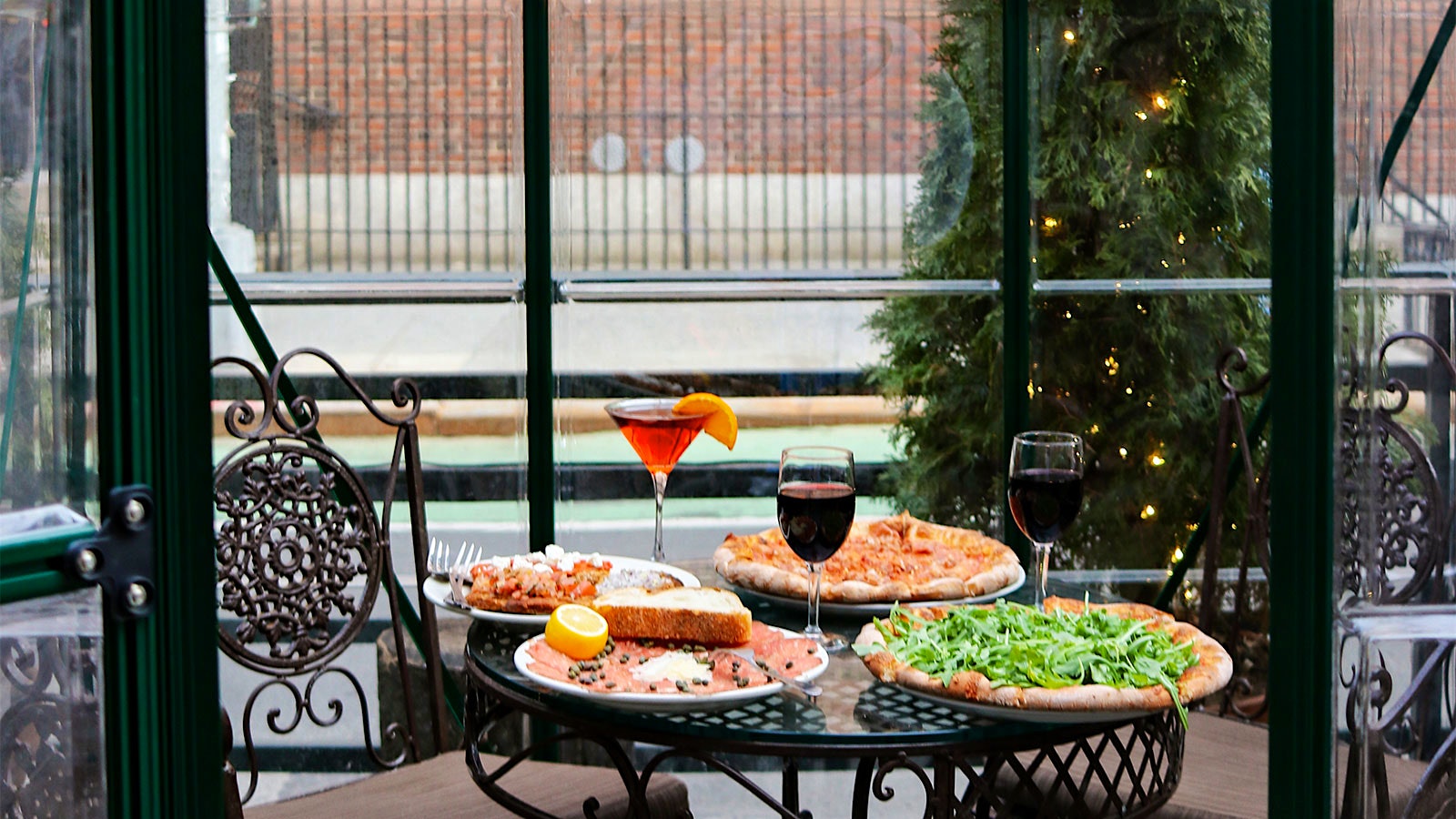 A table within a glass conservatory at La Lanterna di Vittorio, set with a cocktail, two glasses of red wine, two pizzas and smoked salmon